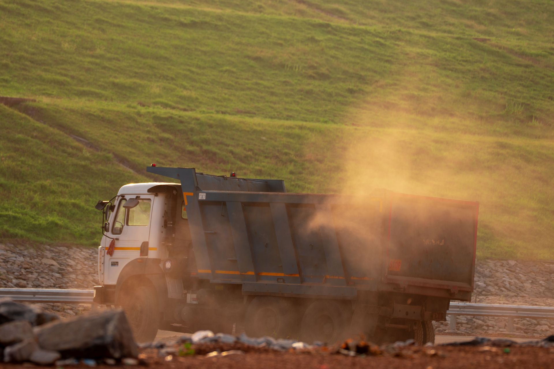A dump truck kicks up dust while driving on a road near grassy hills during daytime.